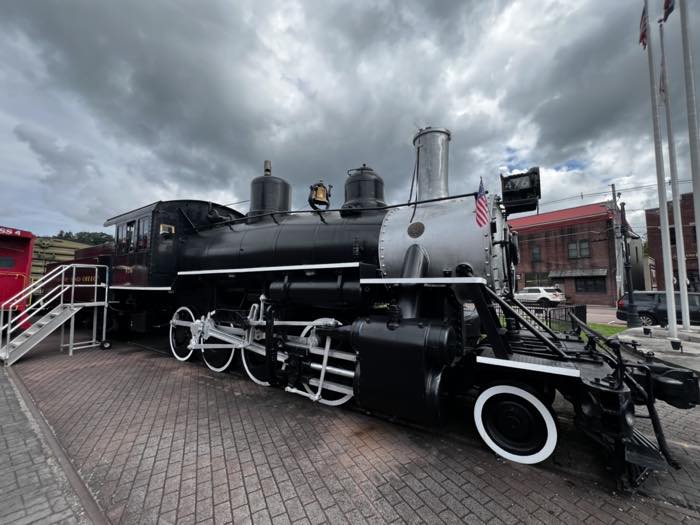 A black steam locomotive numbered 476 with white-rimmed driving wheels, standing on brick paving beside the museum with a red caboose behind it and storm cloud overhead.