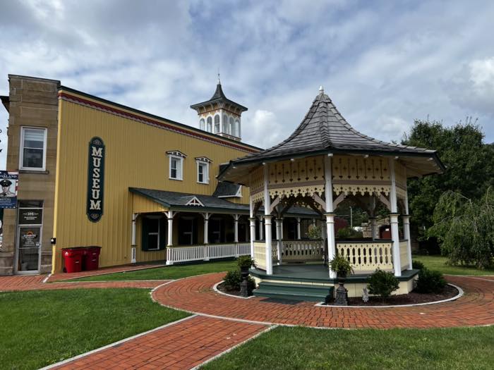 A white and yellow Victorian gazebo with a shingled roof and turned balusters, on a brick apron beside a yellow clapboard building with a tall MUSEUM sign and a cupola.