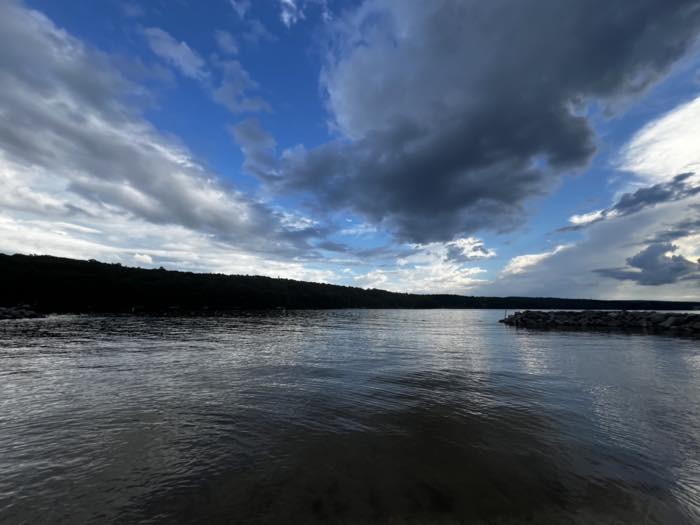 A wide view across the lake from inside the breakwater, with stone piled along both sides of the swim area and a long wooded ridge on the far shore.