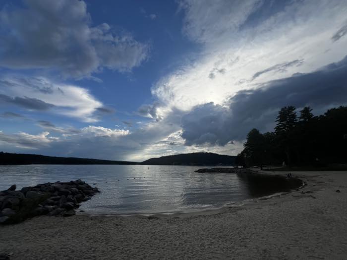 The beach and lake from further along the sand, low sun breaking through heavy cloud over the far hills.