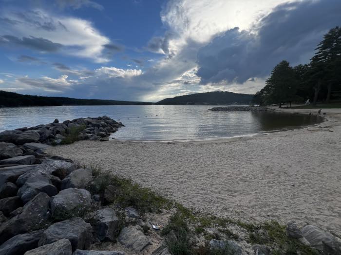 The swimming beach seen along the shore, with a long stone breakwater running out to the left, wooded hills across the lake and pines along the sand.