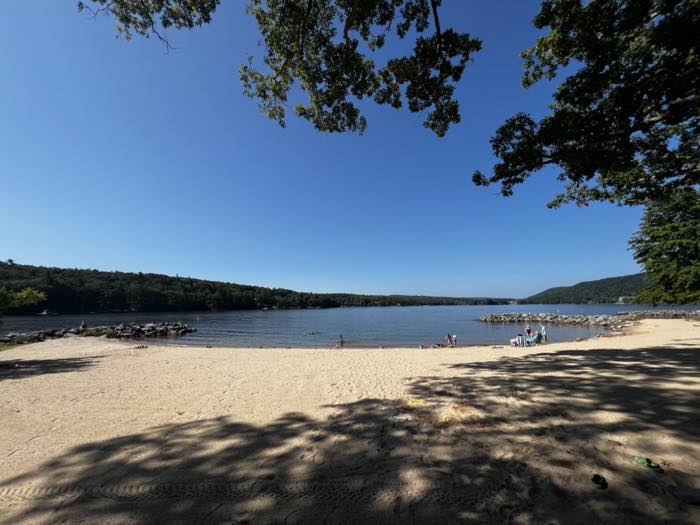 A wide sand beach curving to a lake, with stone breakwaters enclosing a swim area, people swimming and sitting on the sand, and forested hills along the far shore.