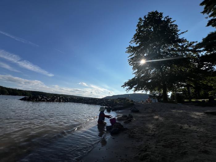 A child in a sun hat crouched at the water's edge with a bucket, on a sand beach inside a long stone breakwater, with wooded hills across the lake and the sun flaring through a big tree.
