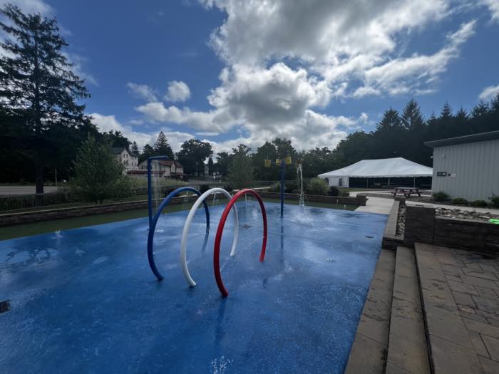 The same splash pad from pad level, with three spray hoops, ground jets, and a tower of yellow tipping buckets, a stone retaining wall and an event tent behind.