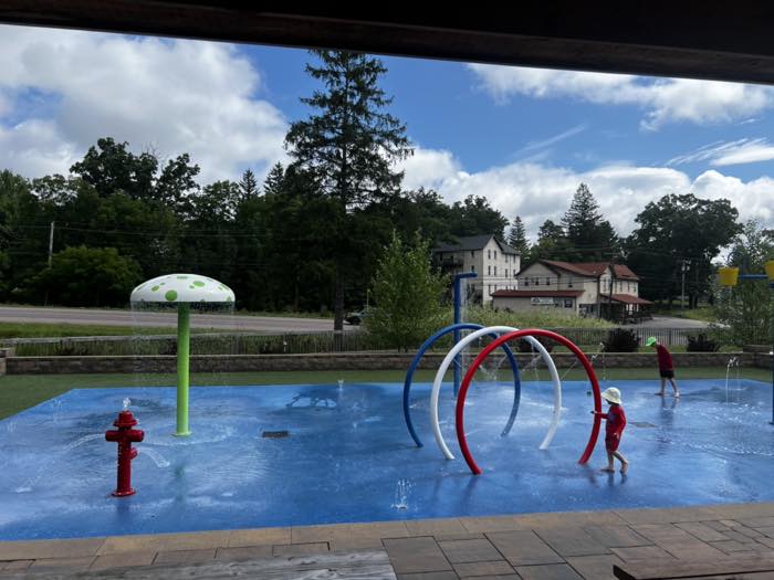 A blue splash pad seen from under a pavilion roof, with a green-and-white mushroom shower, a red fire hydrant spray, three spray hoops in blue, white and red, and two children in sun hats walking through the water.