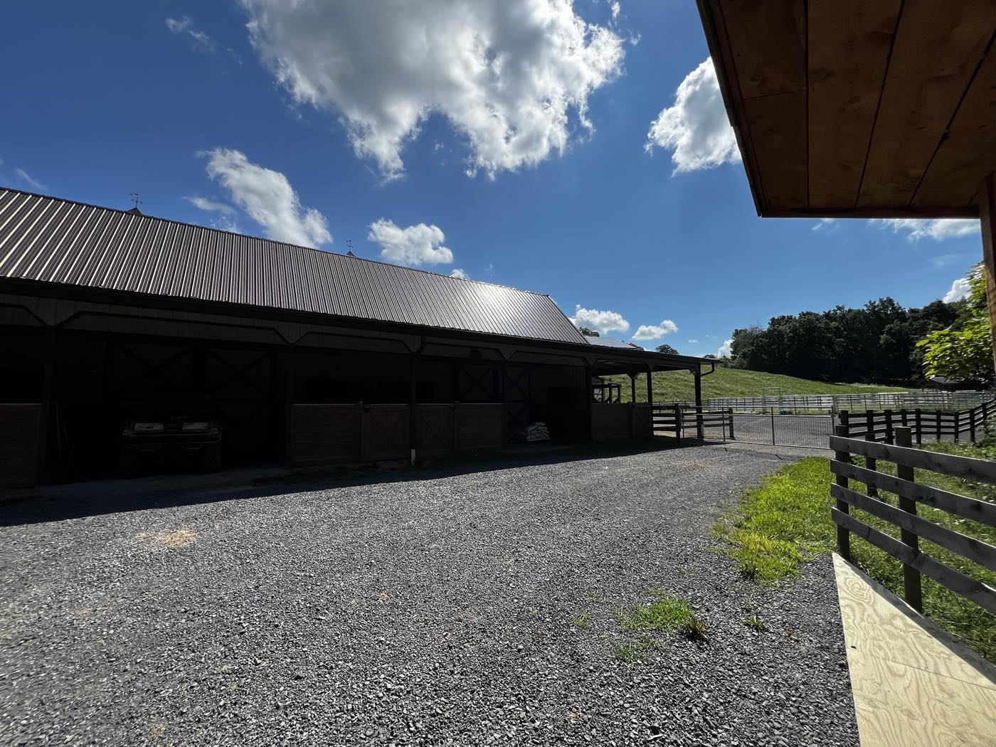 The outside of a long stable block with a brown metal roof, timber dutch doors, a gravel yard and white paddock fencing running up a green hillside.