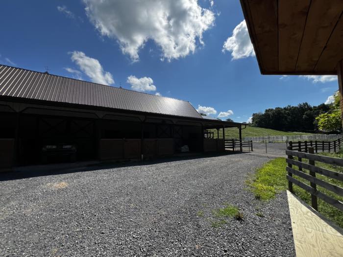 The outside of a long stable block with a brown metal roof, timber dutch doors, a gravel yard and white paddock fencing running up a green hillside.