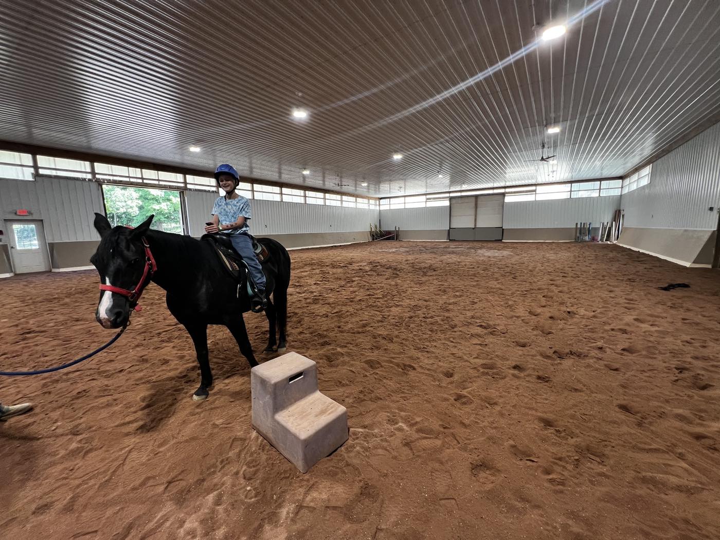 A large indoor riding arena with a sand floor, steel walls, clerestory windows and ceiling fans, with a child in a helmet sitting on a black horse beside a mounting block.