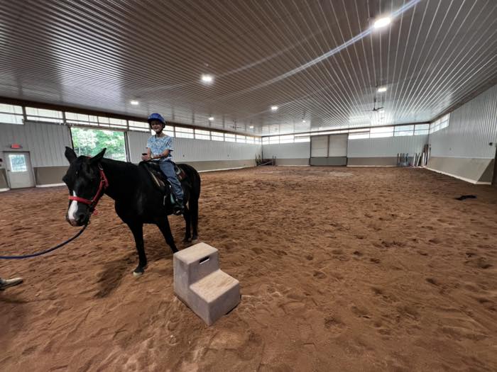 A large indoor riding arena with a sand floor, steel walls, clerestory windows and ceiling fans, with a child in a helmet sitting on a black horse beside a mounting block.
