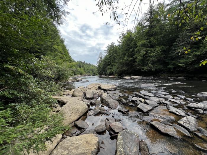 A wide rocky river running between hemlock banks under a cloudy sky, with flat stone shelves and boulders across the near bank.