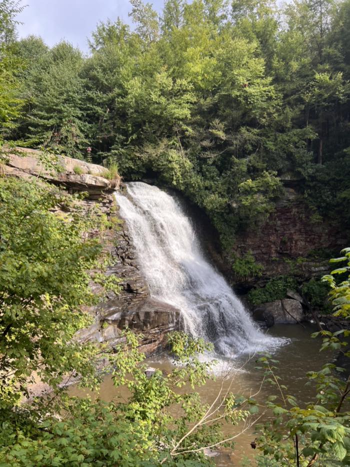 A wide waterfall dropping over a layered rock ledge into a brown pool, framed by summer trees on both sides and a bare rock shelf at the top left.