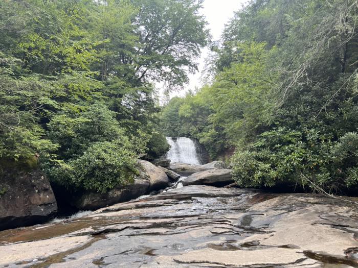 A smaller falls seen from downstream across a broad shelf of flat wet rock, with rhododendron leaning in from both banks and a green canopy above.